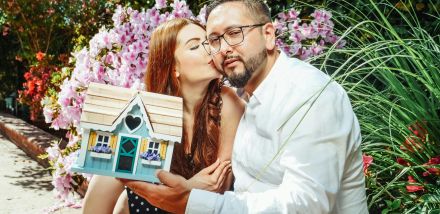 Happy couple with a miniature house model, symbolizing new home purchase, in a vibrant garden.